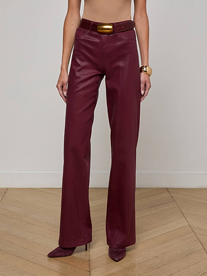 A person stands on a wooden floor wearing L’AGENCE Clayton Coated Wide-Leg Jeans in maroon, matching pointed-toe shoes, a tan sleeveless top, a brown belt with a gold buckle, and a chunky gold bracelet.