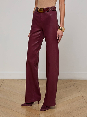 A person wears the L'AGENCE Clayton Coated Wide-Leg Jean in maroon with a matching belt, a tan top, burgundy pointed-toe heels, and a chunky gold bracelet, standing on a wooden floor against a plain wall.