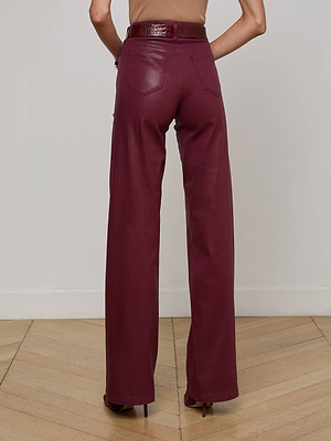A person stands with their back to the camera, wearing L'AGENCE Clayton Coated Wide-Leg Jean in maroon with a brown belt, on a wooden floor and white wall background.