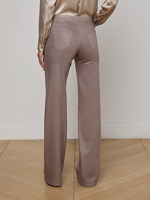 A person wearing L'AGENCE's Clayton Coated Wide-Leg Jean stands on a wooden floor, facing away from the camera with their left hand on their hip, paired with a silky beige shirt.