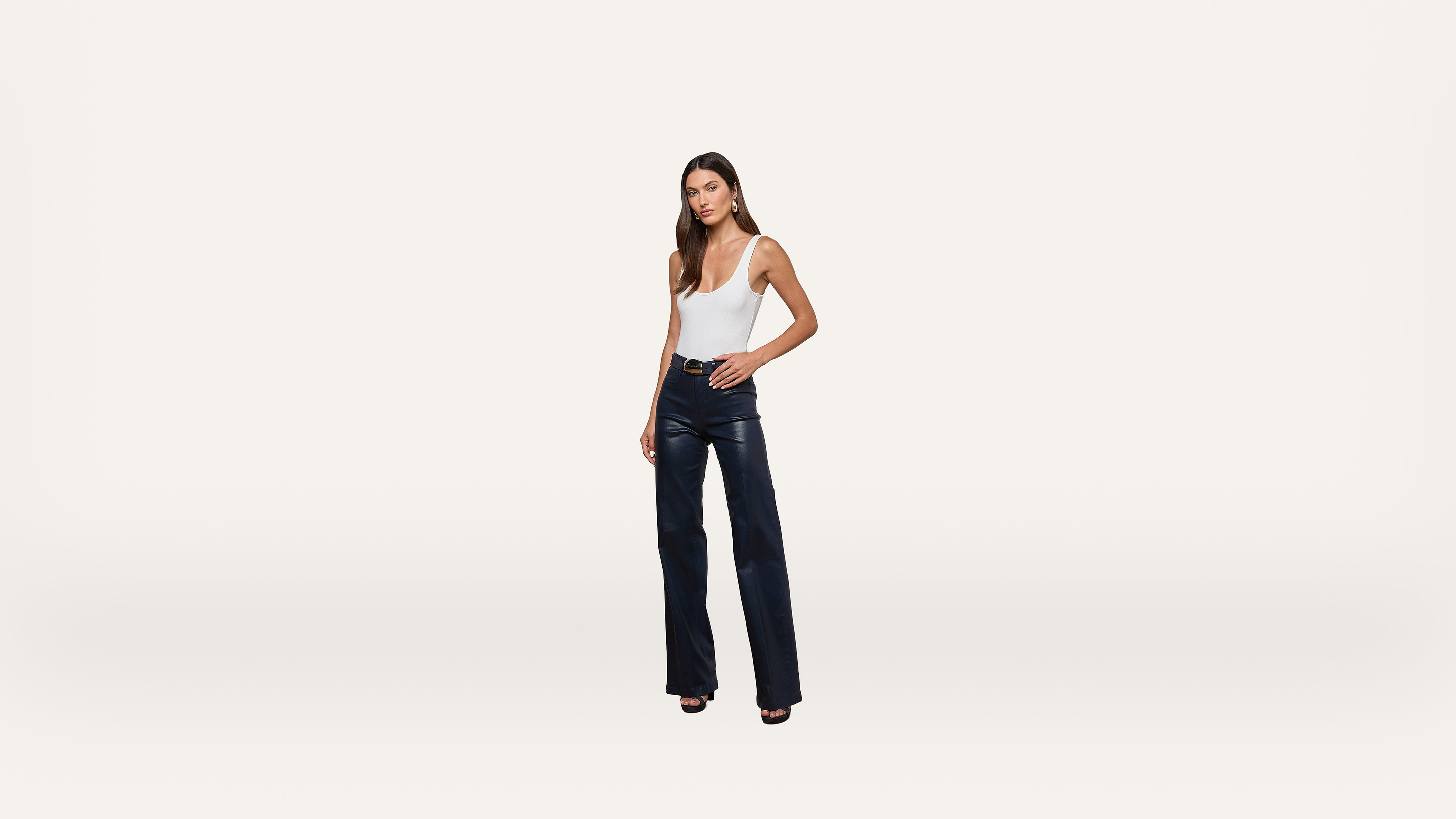A woman stands against a plain white background, wearing a white sleeveless top, black leather pants, and black open-toe heels.