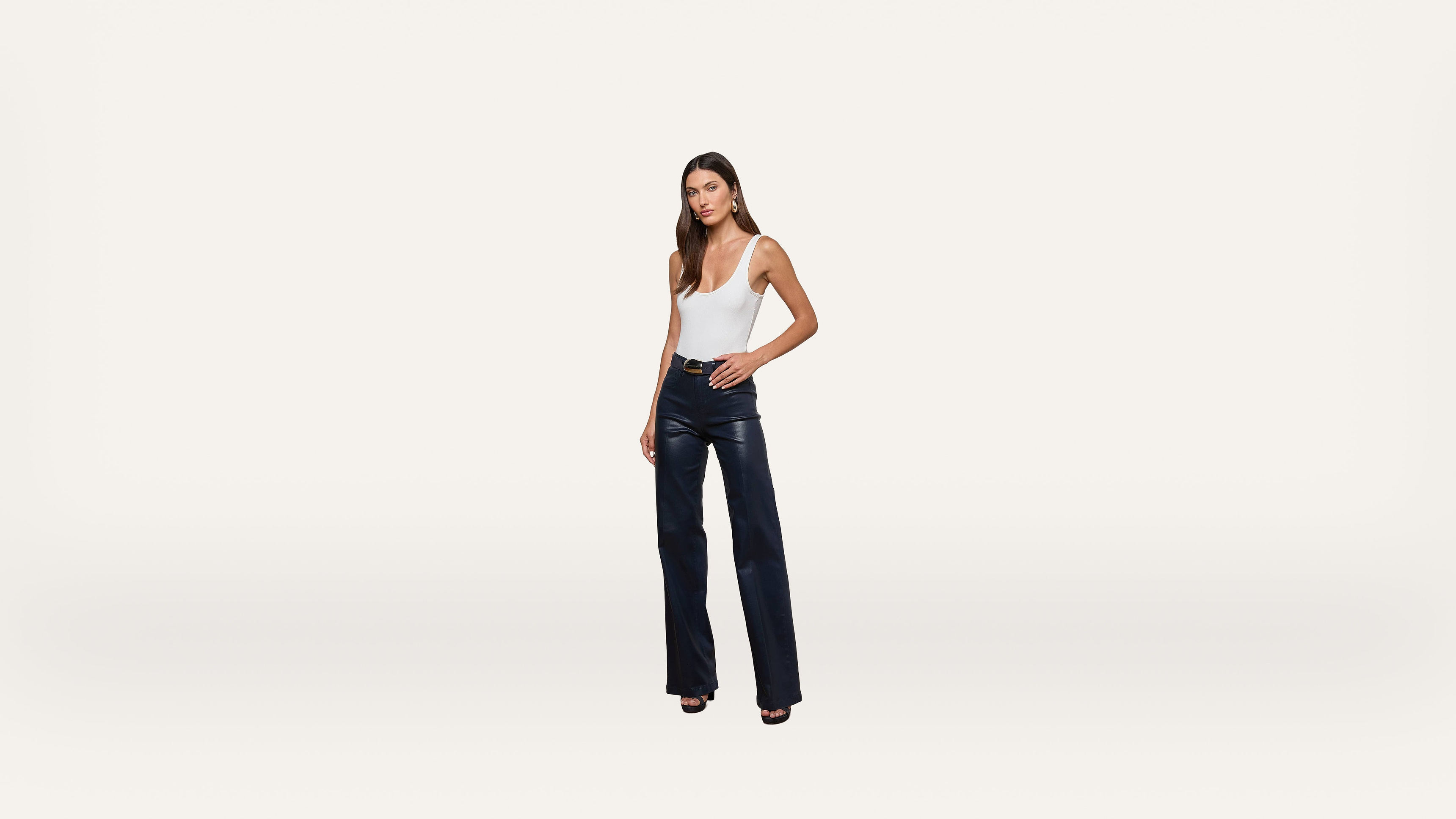 A woman stands against a plain white background, wearing a white sleeveless top, black leather pants, and black open-toe heels.