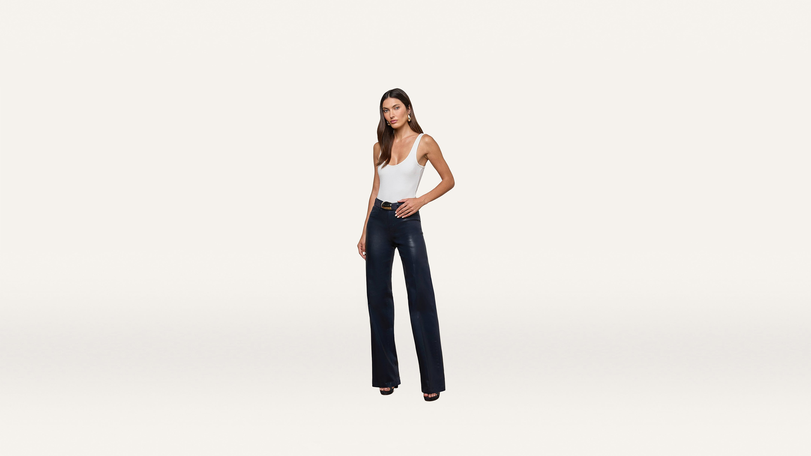 A woman stands facing forward, wearing a white sleeveless top and high-waisted dark flared pants with black heels, styled with a modern and minimalist touch against a plain white background.