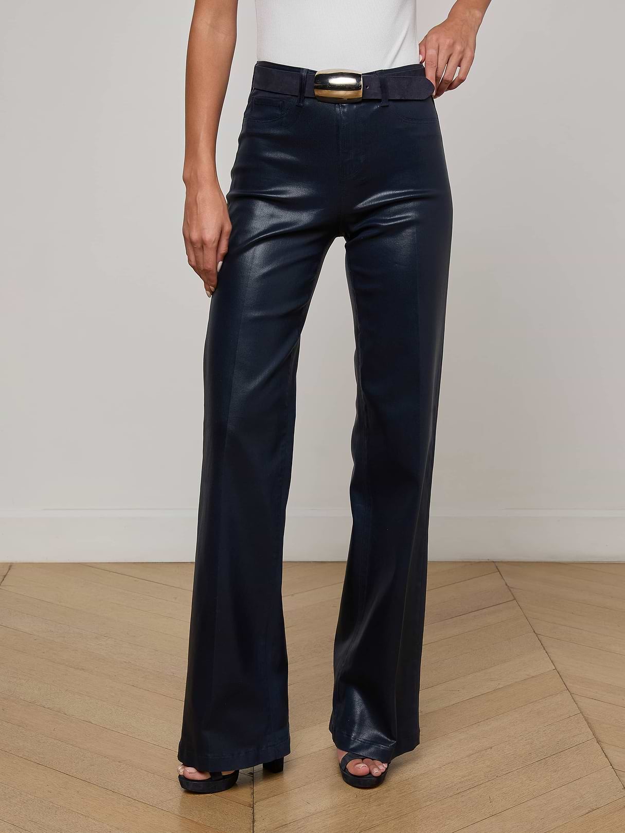 A person is shown from the waist down, wearing the Clayton Coated Wide-Leg Jean in black paired with a black belt and a white top, standing against a plain background.