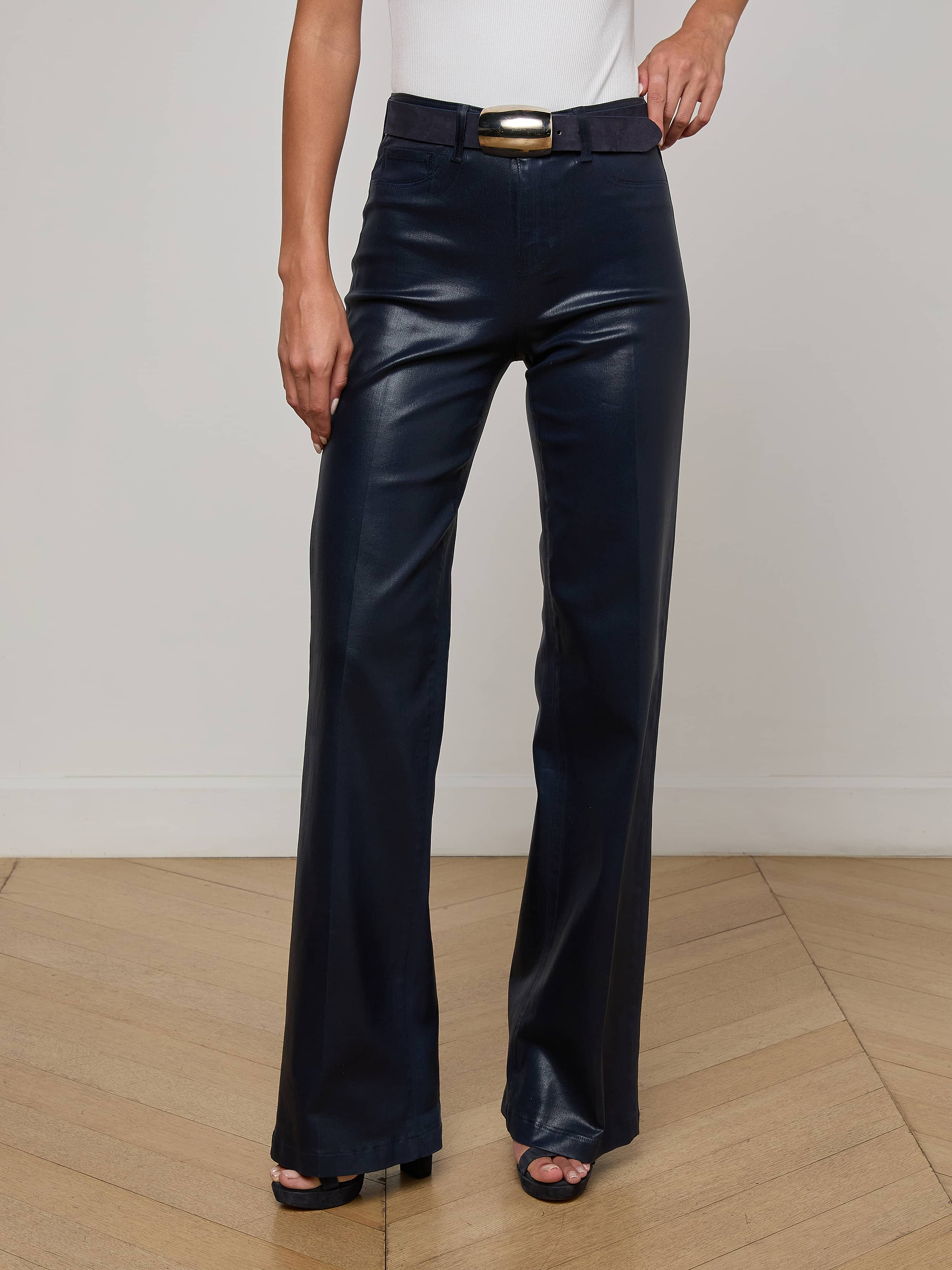 Wearing the L'AGENCE Clayton Coated Wide-Leg Jean with a matching belt and a white sleeveless top, a person stands on a wooden floor with one hand relaxed by their side.