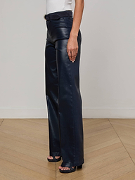 Wearing the L'AGENCE Clayton Coated Wide-Leg Jean and black open-toe heels, a person stands on a light wood floor against a plain white wall with one arm relaxed at their side.