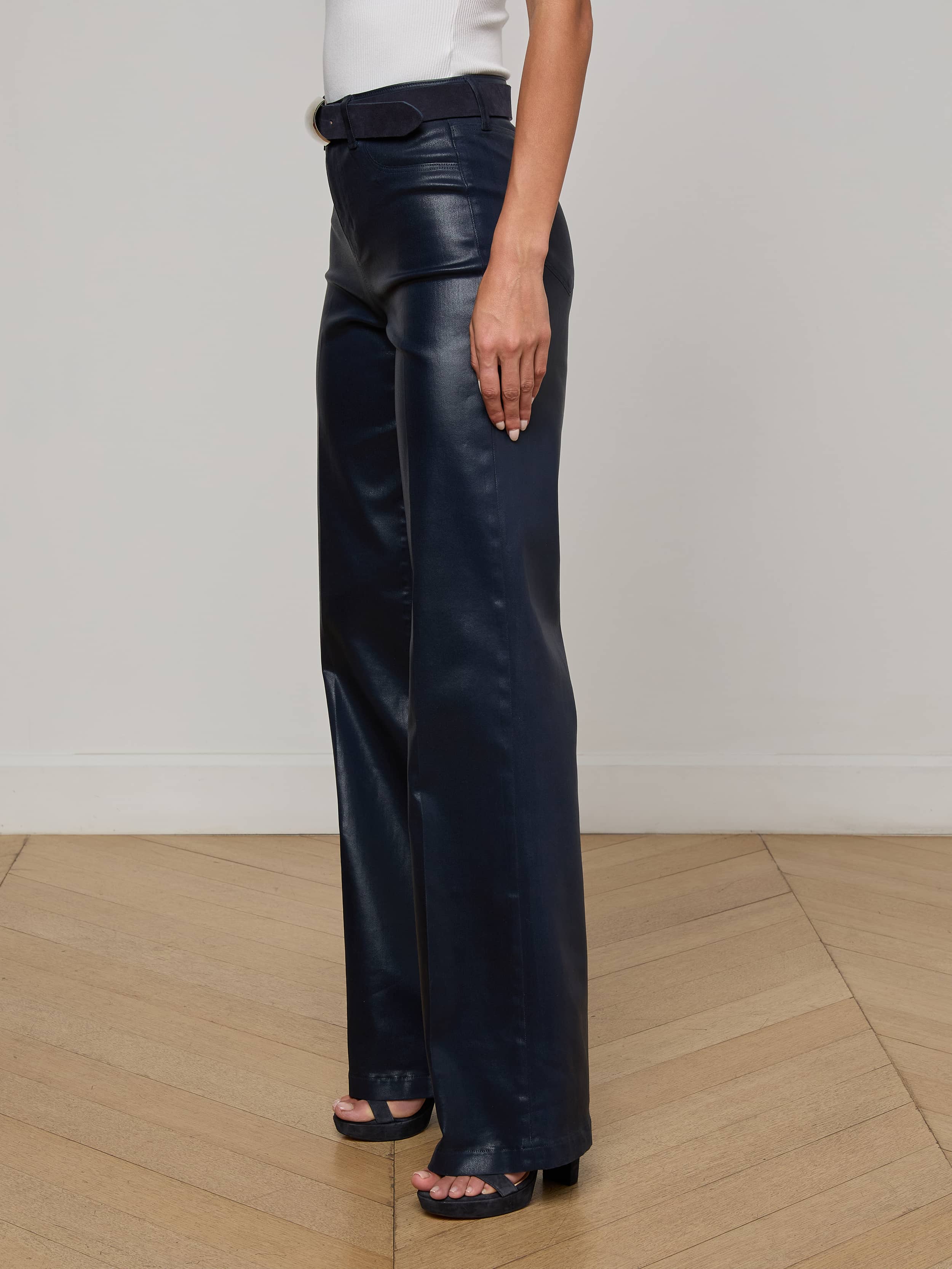 Wearing the L'AGENCE Clayton Coated Wide-Leg Jean and black open-toe heels, a person stands on a light wood floor against a plain white wall with one arm relaxed at their side.