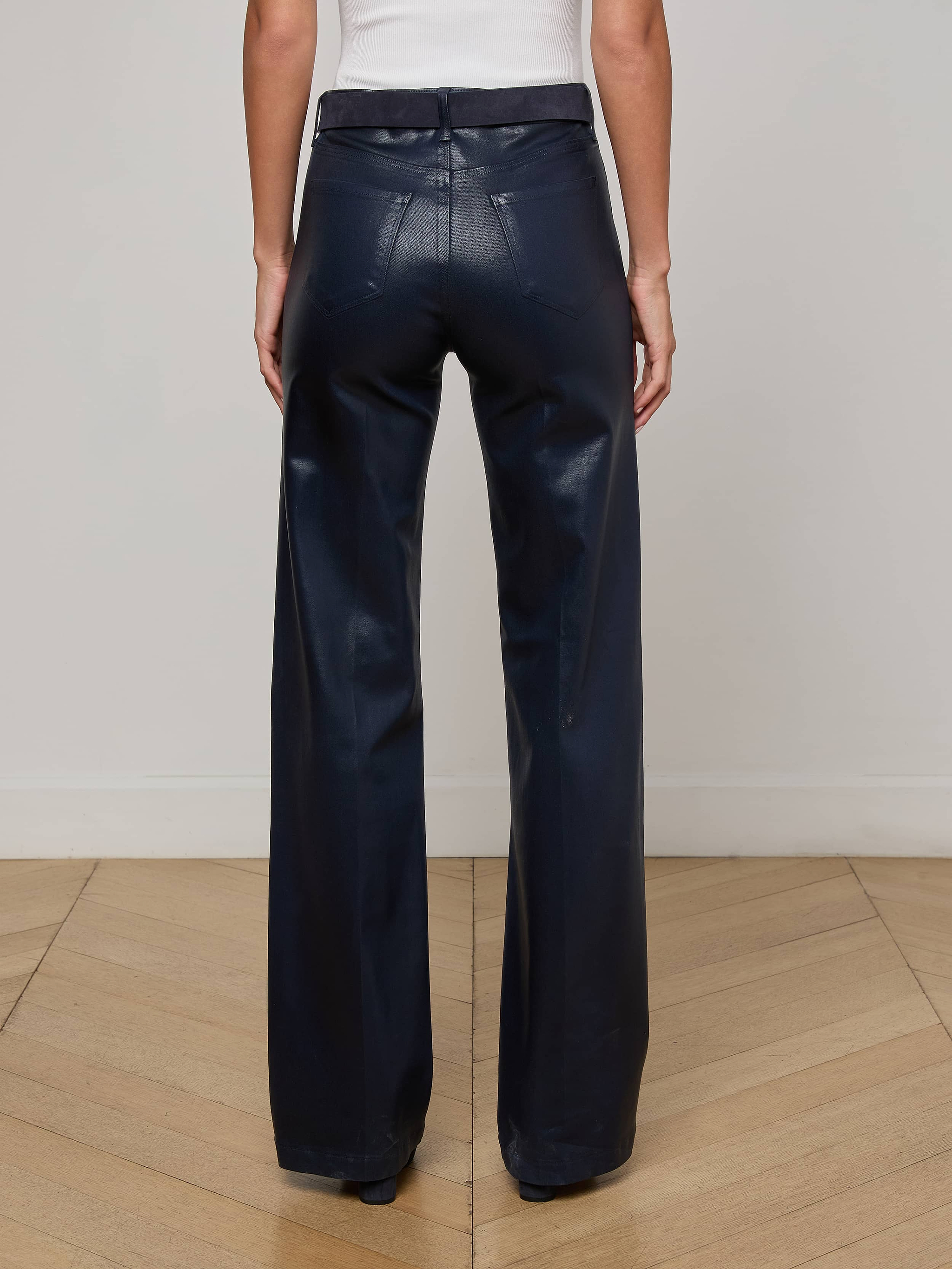 A person stands on a wooden floor, back to the camera, showcasing the L'AGENCE Clayton Coated Wide-Leg Jean in dark navy with a sleeveless white top.