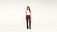 A woman stands facing forward, wearing a sleeveless white top and high-waisted brown leather pants with a belt, against a plain white background.