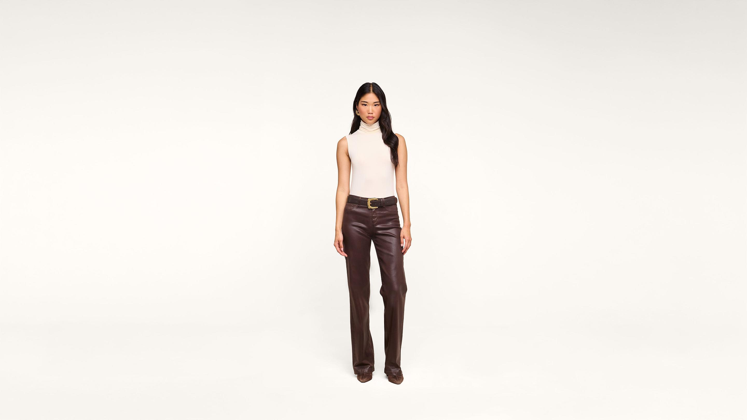 A woman stands facing forward, wearing a sleeveless white top and high-waisted brown leather pants with a belt, against a plain white background.