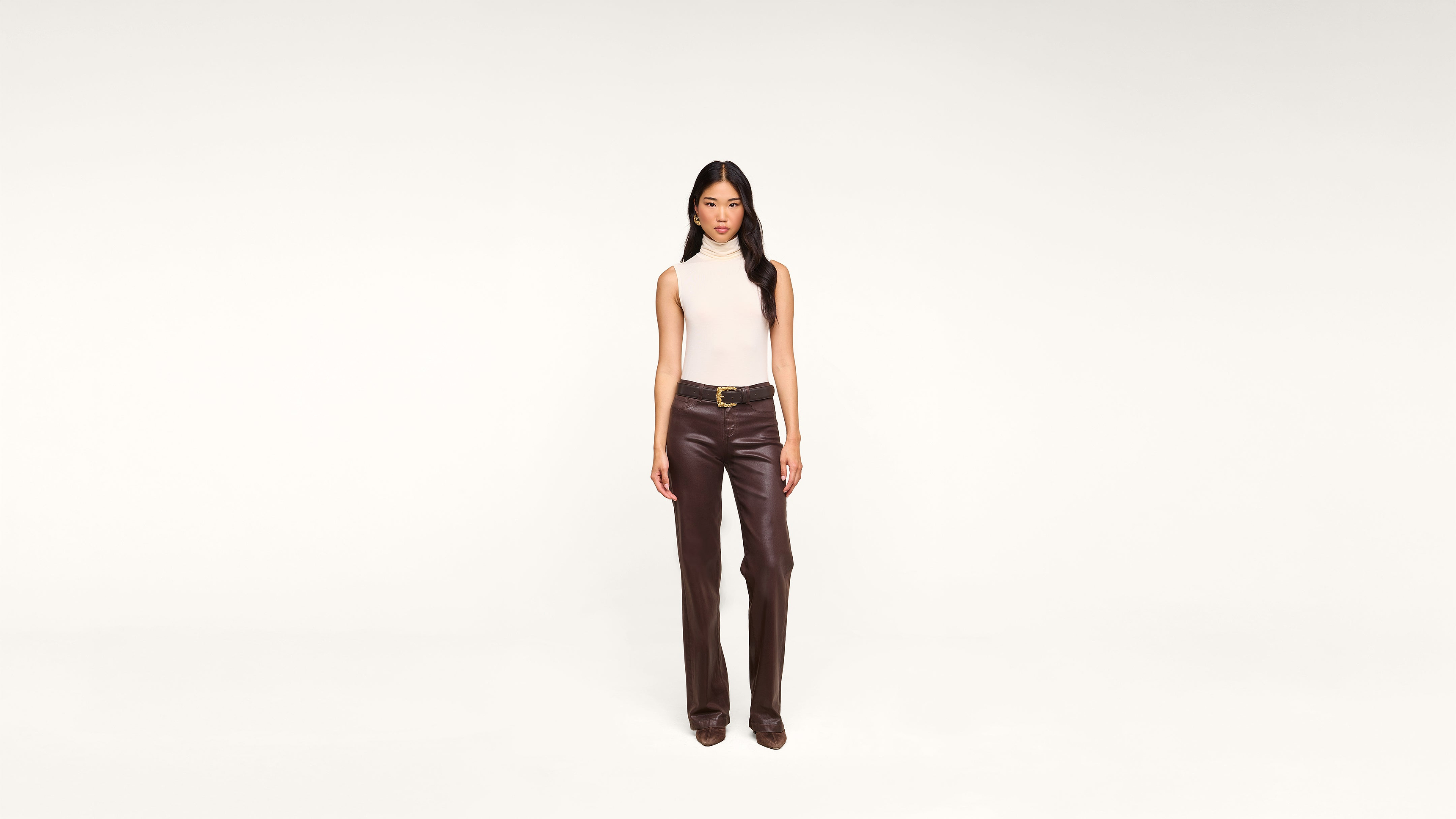 A woman stands facing forward, wearing a sleeveless white top and high-waisted brown leather pants with a belt, against a plain white background.