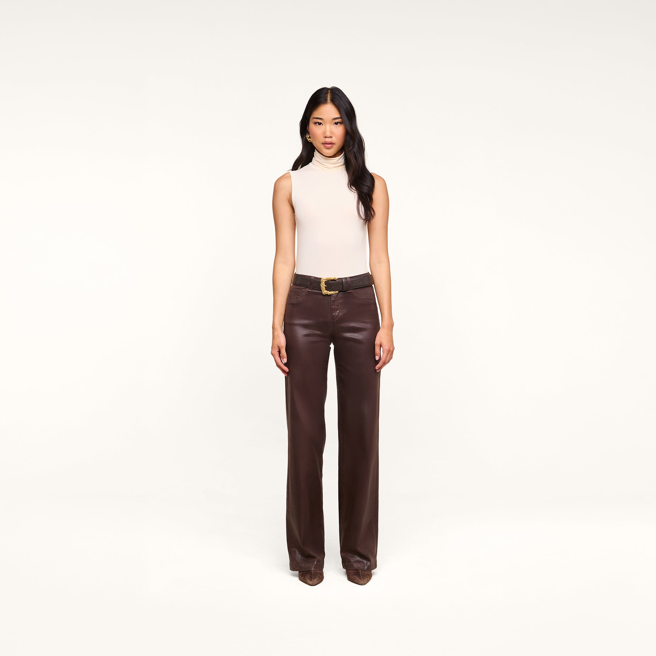 A woman stands facing forward against a plain white background, wearing a sleeveless beige top, brown shoes, and the Clayton Coated Wide-Leg Jean—dark brown high-rise coated denim pants with a belt.