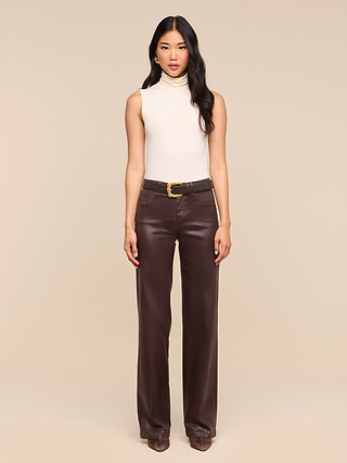 A woman stands facing forward against a plain beige background, wearing a sleeveless white turtleneck and the Clayton Coated Wide-Leg Jean, styled with a black belt and brown shoes.