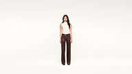 A woman stands against a plain white background, wearing a sleeveless beige top and high-waisted dark brown pants with a belt. She faces forward with a neutral expression, her chic ensemble highlighting timeless minimalist style.