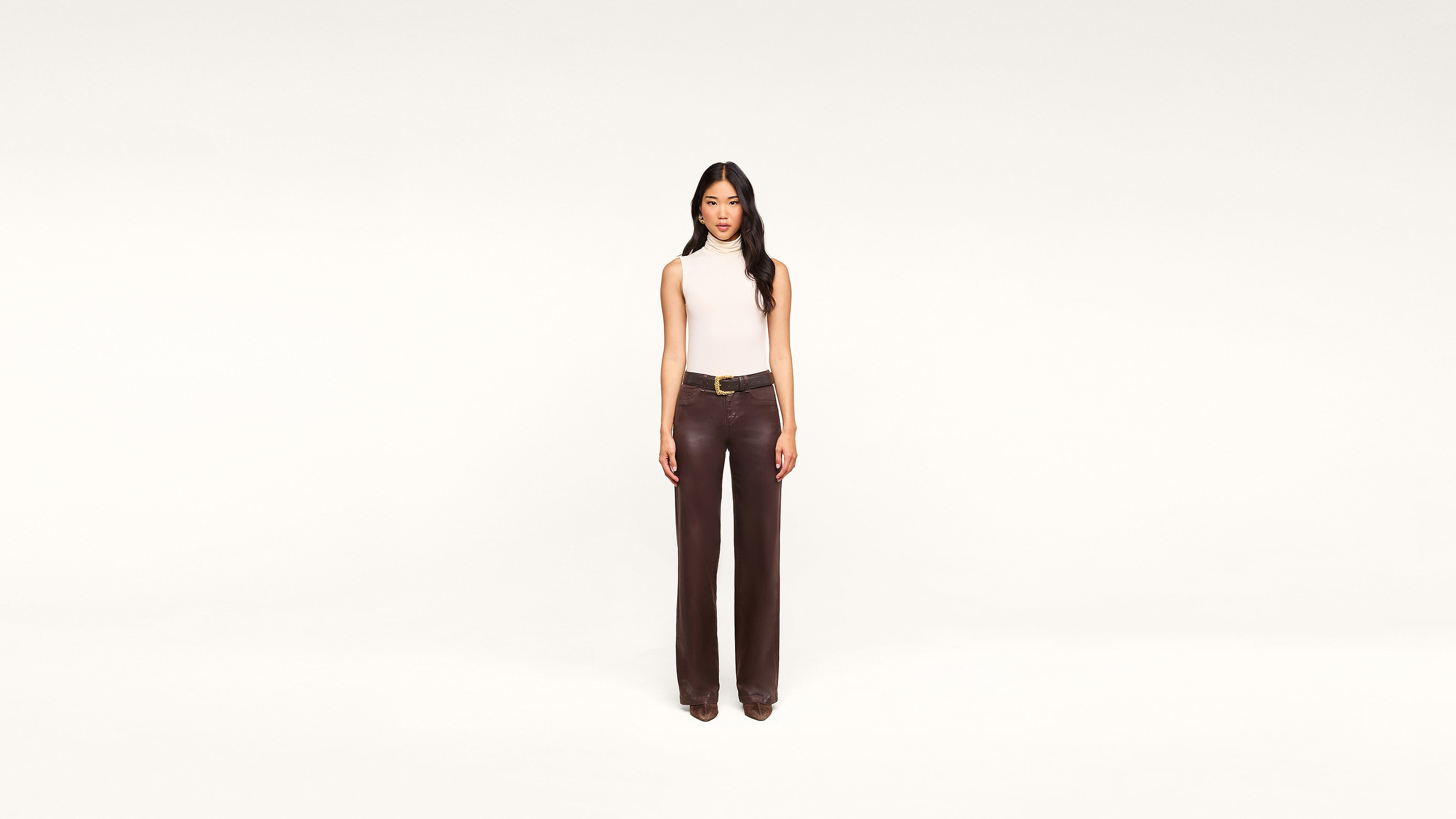 A woman stands against a plain white background, wearing a sleeveless beige top and high-waisted dark brown pants with a belt. She faces forward with a neutral expression, her chic ensemble highlighting timeless minimalist style.