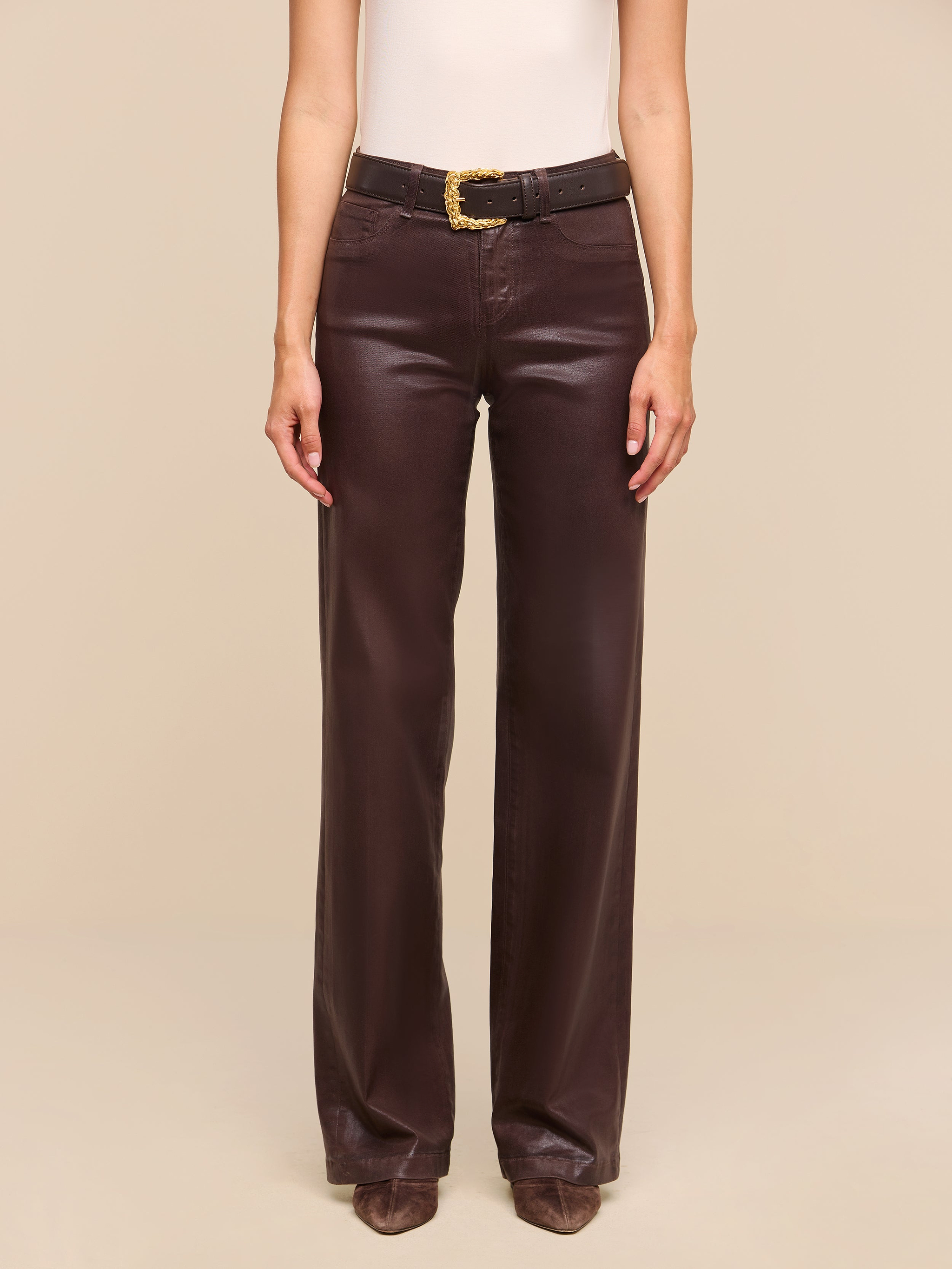 A person is standing against a plain white background, wearing a sleeveless beige top, the Clayton Coated Wide-Leg Jean in dark brown with a black belt, and brown pointed shoes.