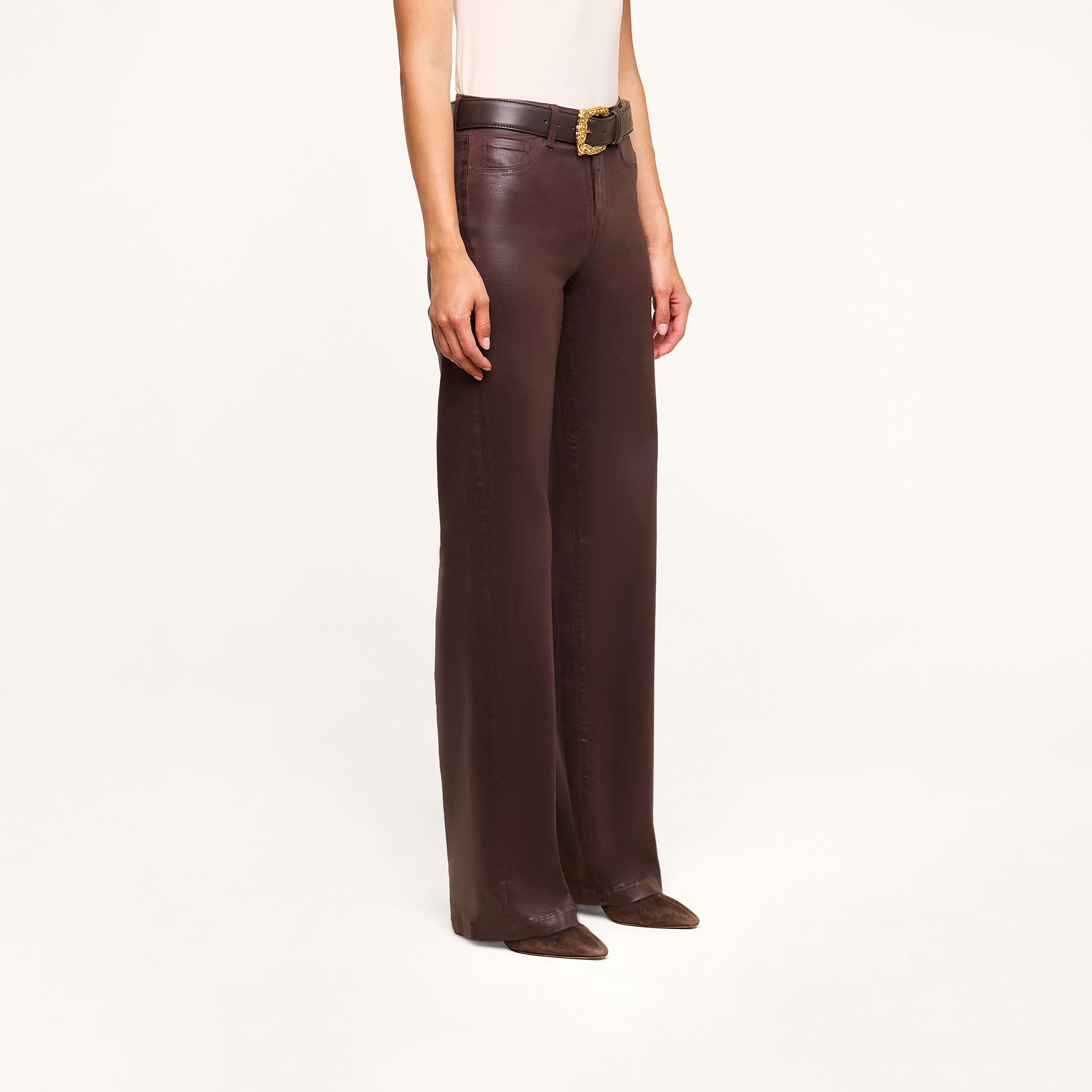 A person wearing Clayton Coated Wide-Leg Jeans with a black belt and pointed brown shoes stands against a plain white background.