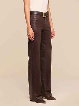A person wearing the Clayton Coated Wide-Leg Jean in dark brown, styled with a black belt featuring a decorative gold buckle and pointed brown shoes, stands against a beige background.