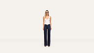 A woman in sunglasses, a white tank top, dark high-waisted wide-leg pants, and sandals stands confidently against a plain white background.