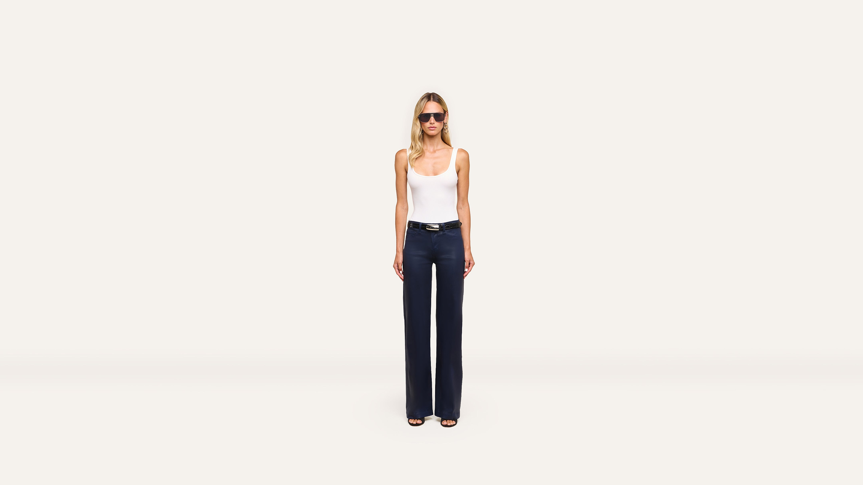 A woman in sunglasses, a white tank top, dark high-waisted wide-leg pants, and sandals stands confidently against a plain white background.
