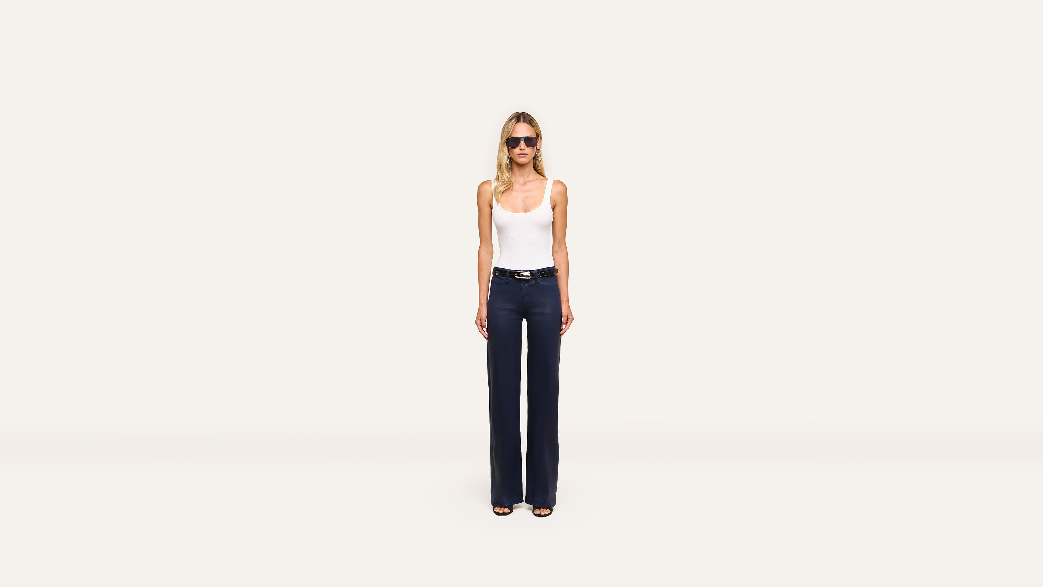 A woman in sunglasses, a white tank top, dark high-waisted wide-leg pants, and sandals stands confidently against a plain white background.