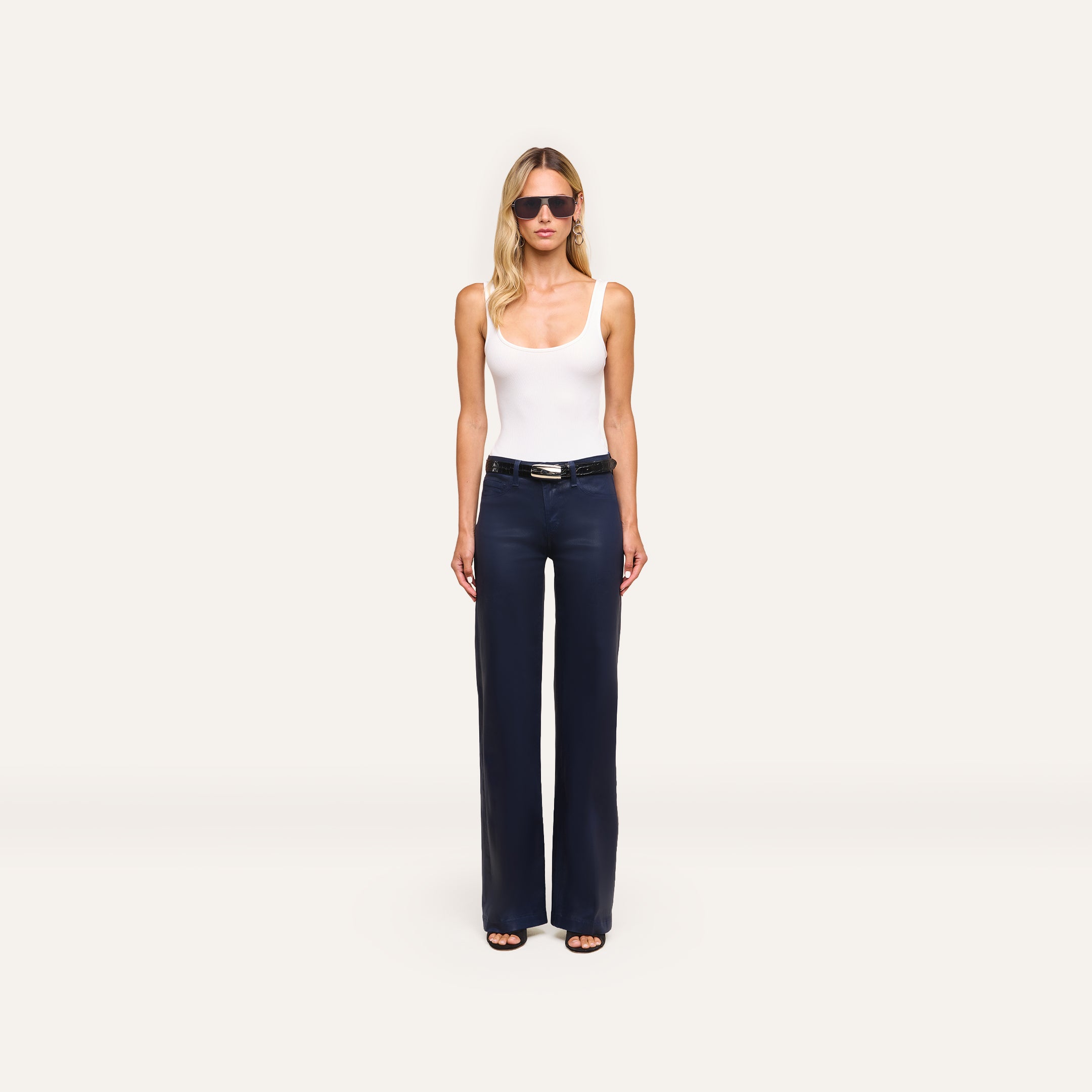 A woman wearing sunglasses, a white tank top, black belt, and Clayton Coated Wide-Leg Jean stands facing forward against a plain light background.