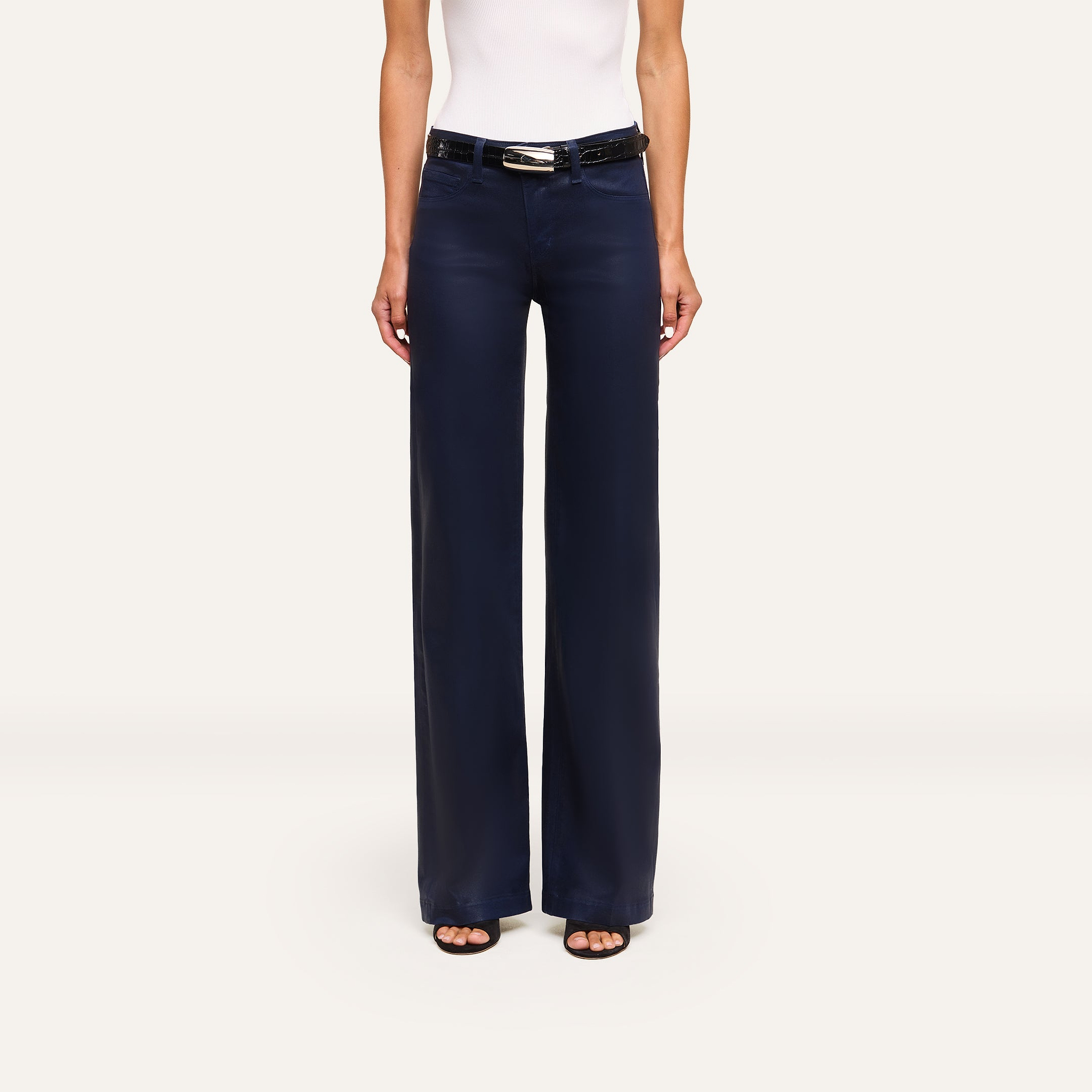 A person wearing the Clayton Coated Wide-Leg Jean in navy blue, a black belt, a white sleeveless top, and black open-toe shoes faces forward against a plain light background.