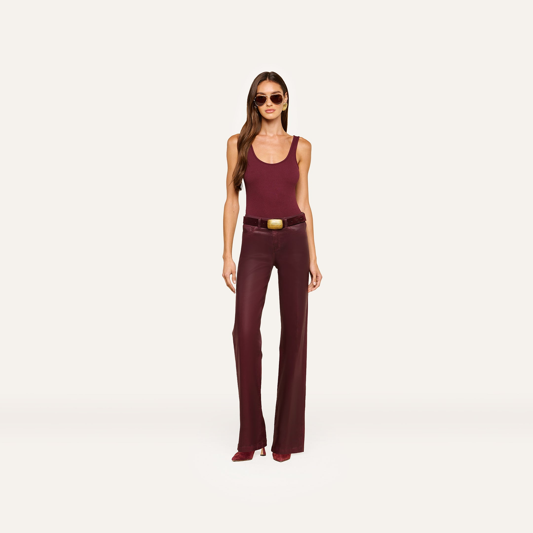 A woman wearing sunglasses, a sleeveless maroon top, the Clayton Coated Wide-Leg Jean with a contoured waistband and belt in dark maroon, and maroon heeled shoes, stands against a plain light background.