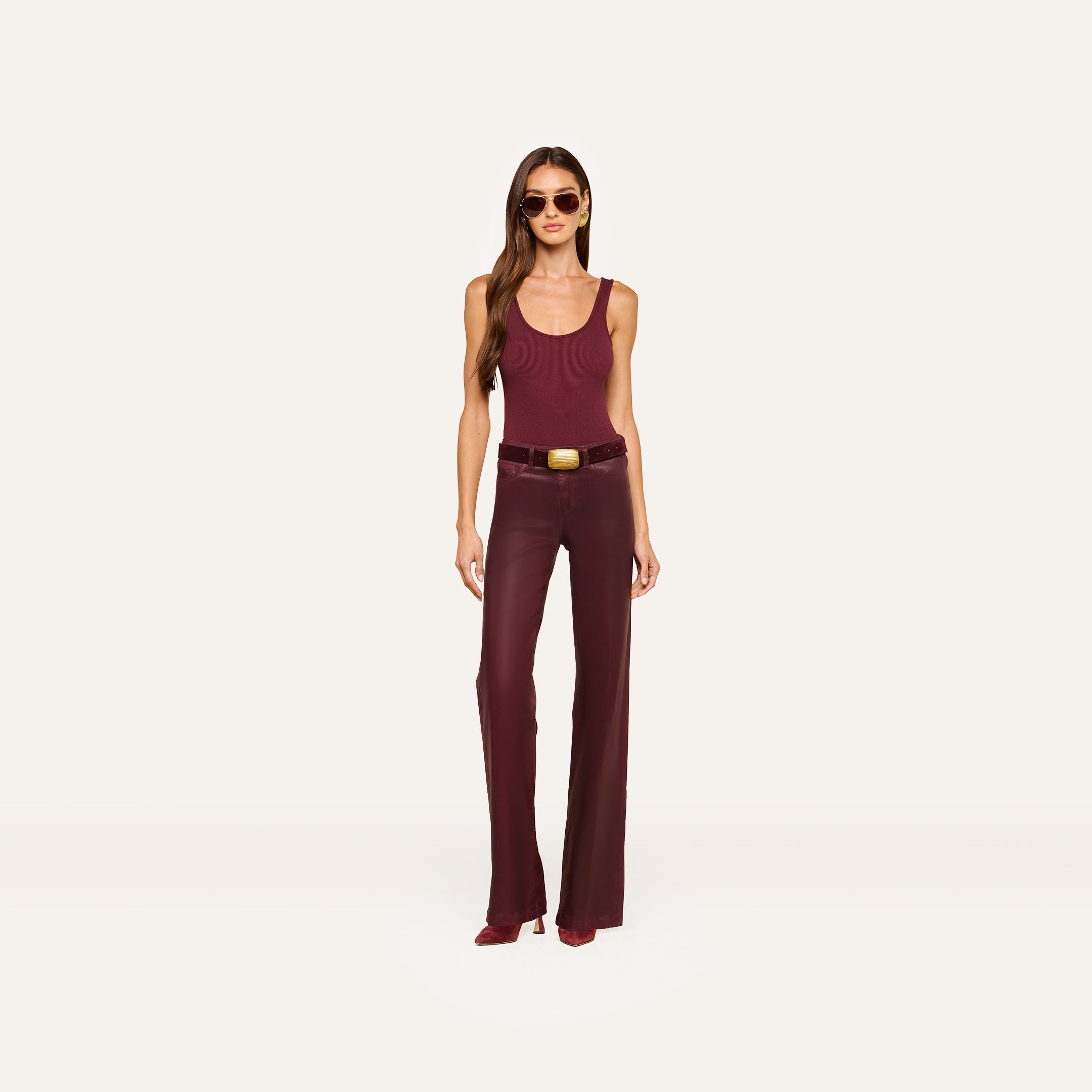 A woman wearing sunglasses, a sleeveless maroon top, the Clayton Coated Wide-Leg Jean with a contoured waistband and belt in dark maroon, and maroon heeled shoes, stands against a plain light background.