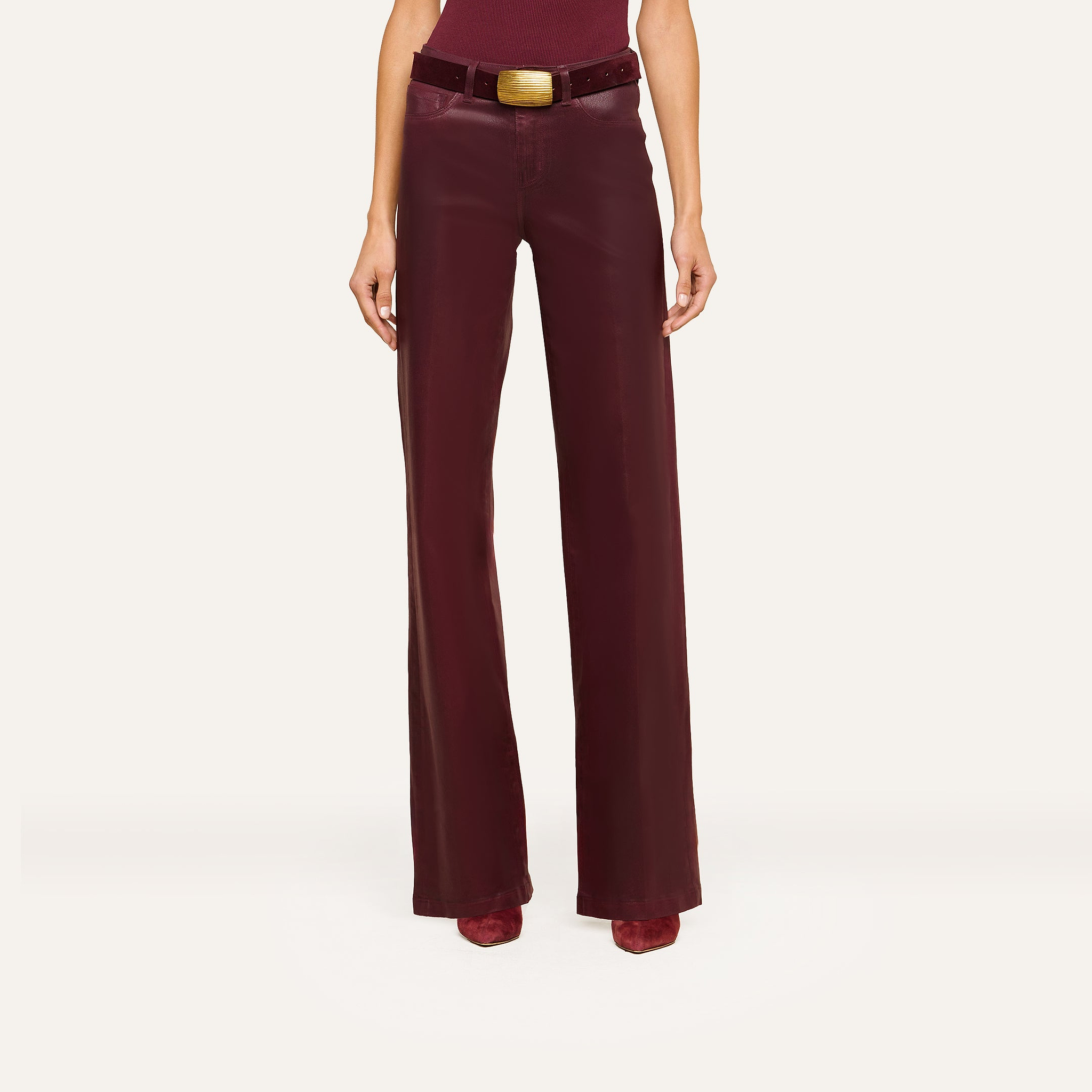 A person wearing high-waisted burgundy leather pants, a burgundy top, matching boots, and a wide belt with a large gold buckle stands confidently against a plain white background.