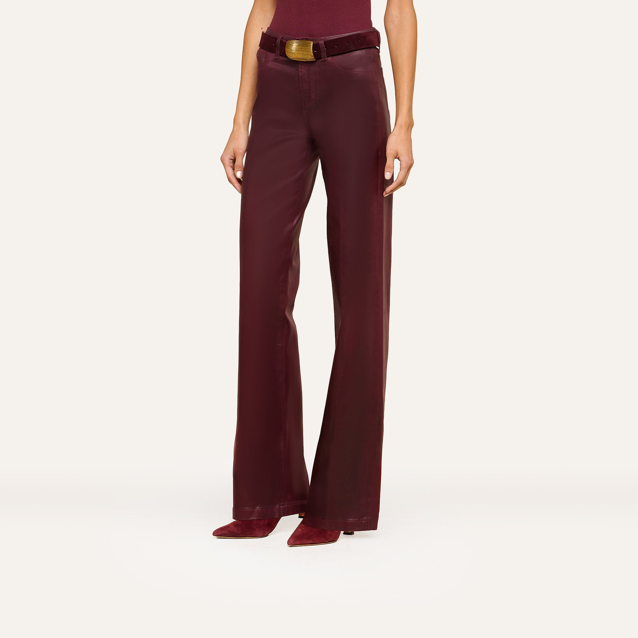 A person models the Clayton Coated Wide-Leg Jean, featuring a high waist, wide-leg silhouette, contoured waistband, and matching belt in burgundy leather. They pair the jeans with pointed burgundy heeled shoes. The top of their head is not visible.