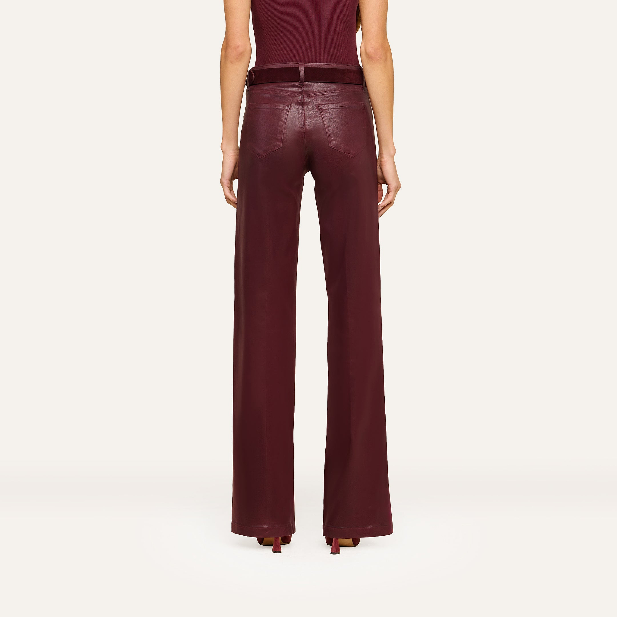 A person stands facing away, wearing the Clayton Coated Wide-Leg Jean featuring a contoured waistband in a rich burgundy hue, paired with a matching belt and heels. The background is plain and light-colored.