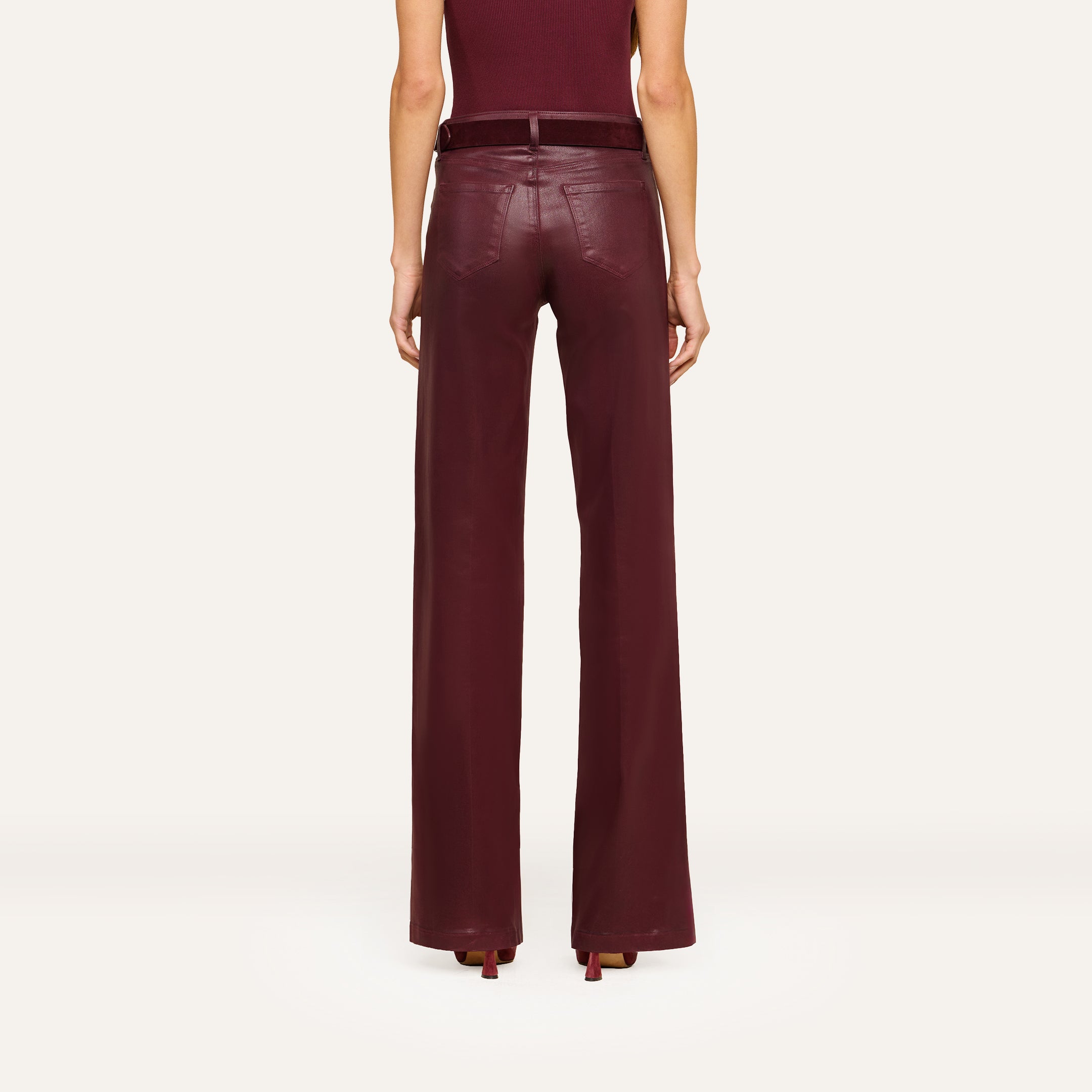 A person stands facing away, wearing the Clayton Coated Wide-Leg Jean featuring a contoured waistband in a rich burgundy hue, paired with a matching belt and heels. The background is plain and light-colored.