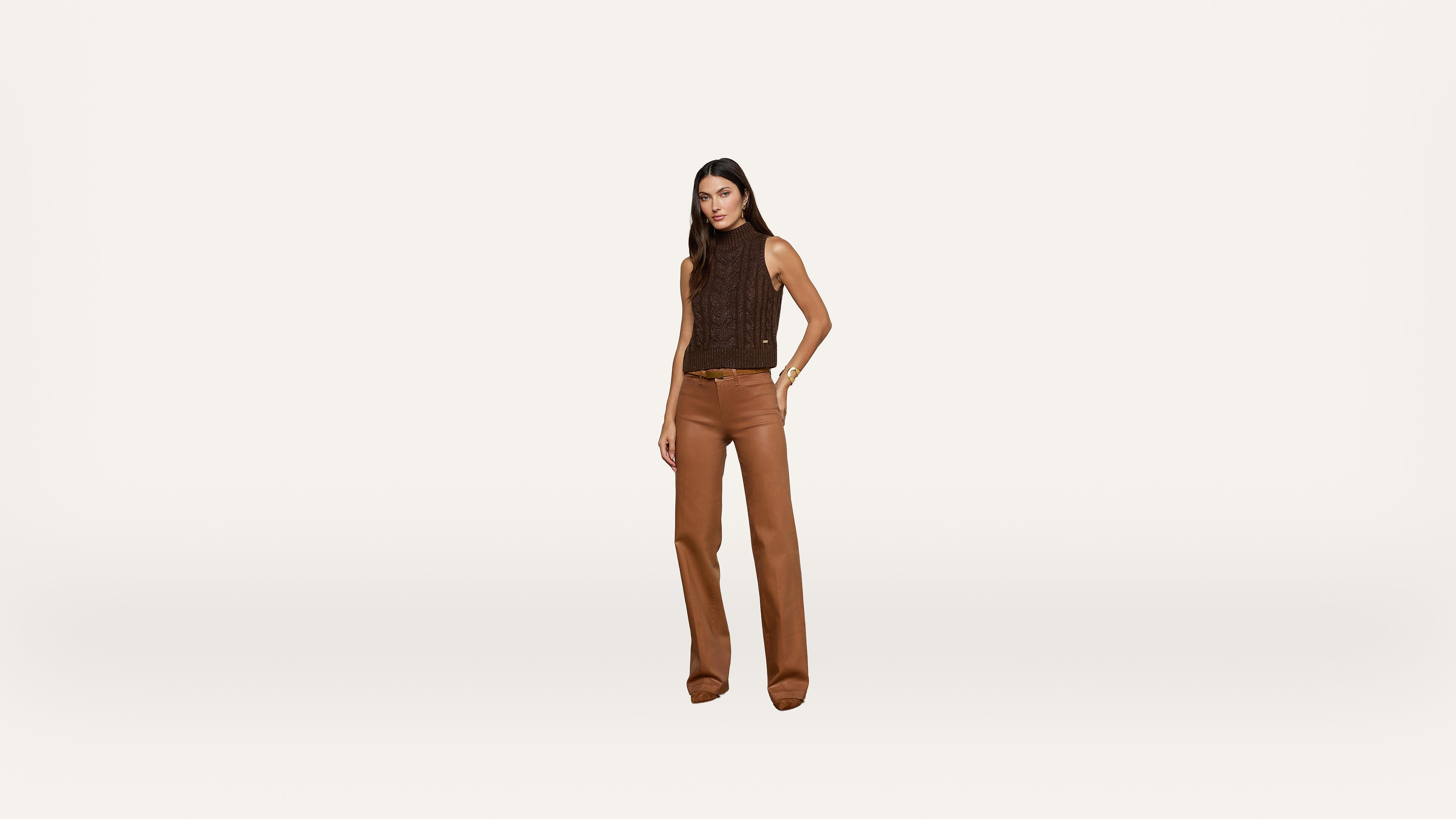 A woman stands against a plain white background wearing a sleeveless brown top and brown wide-leg pants, with one hand in her pocket.