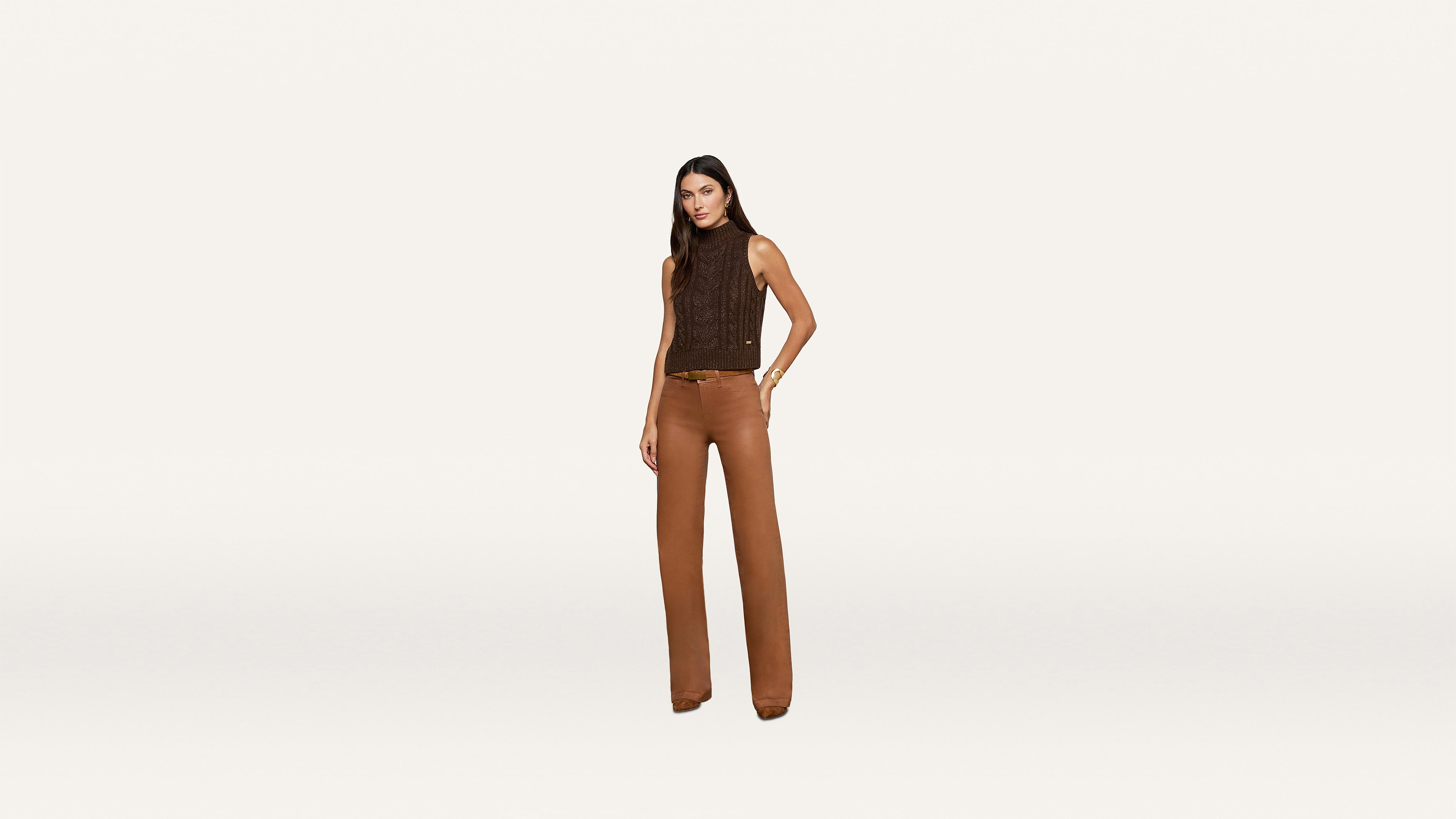 A woman stands wearing a sleeveless brown top and high-waisted brown pants against a plain white background, showcasing a chic minimalist style.
