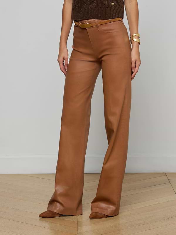 A person stands against a light wall wearing the Clayton Coated Wide-Leg Jean by L'AGENCE, styled with a contoured waistband, matching belt, brown pointed shoes, a sleeveless brown top, and a chunky gold bracelet.
