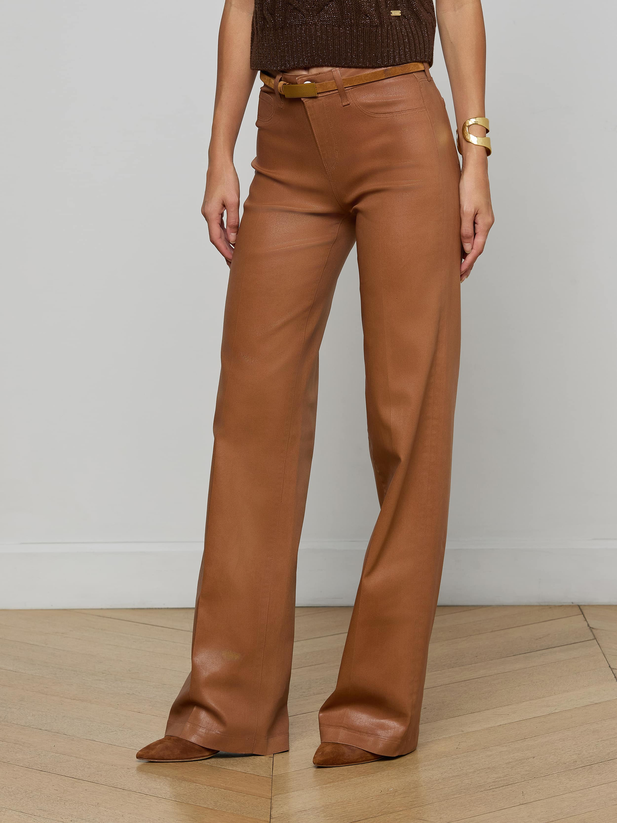 A person stands against a light wall wearing the Clayton Coated Wide-Leg Jean by L'AGENCE, styled with a contoured waistband, matching belt, brown pointed shoes, a sleeveless brown top, and a chunky gold bracelet.