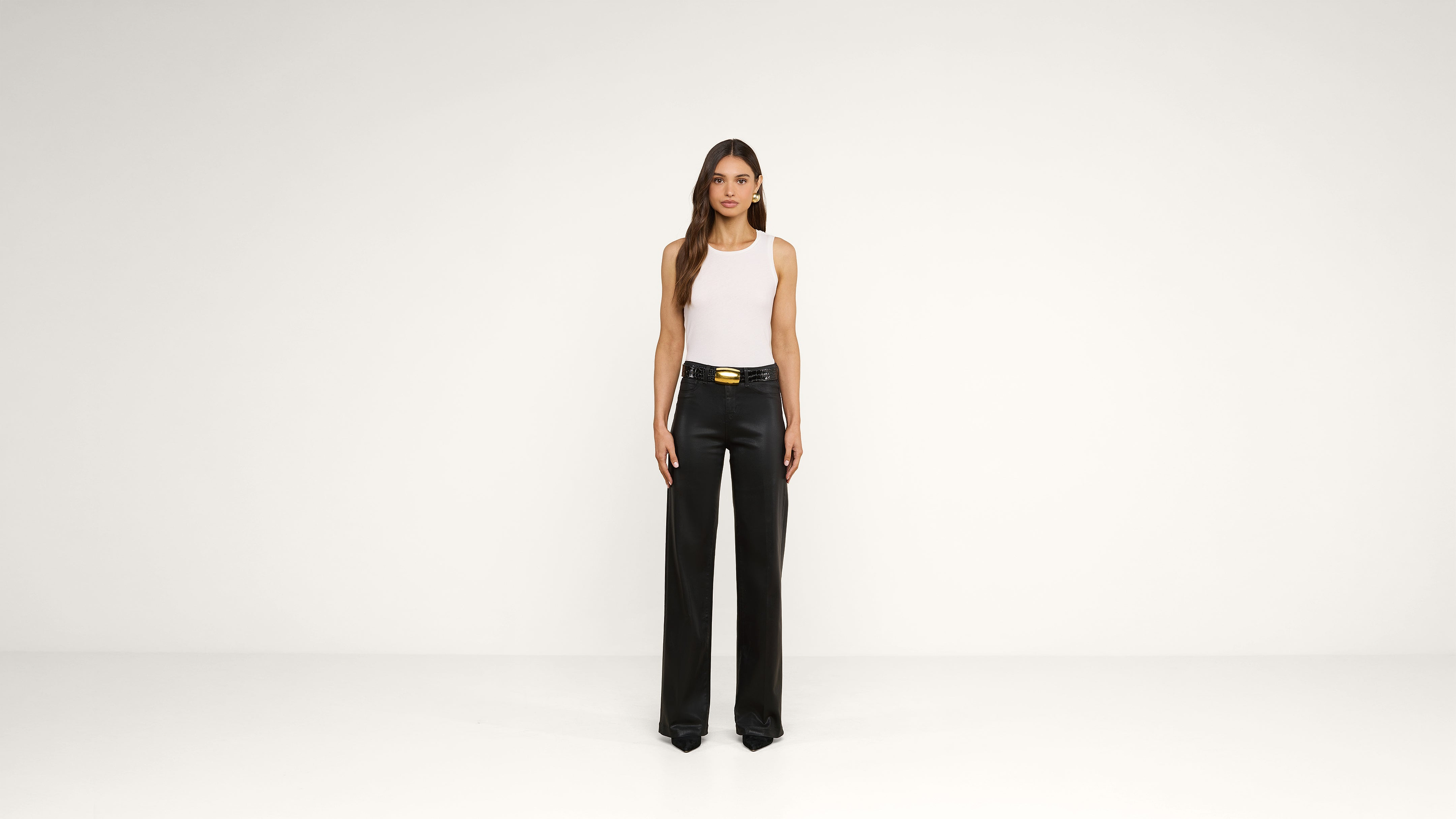 A woman stands against a plain white background, wearing a white sleeveless top, black wide-leg pants with a gold belt buckle, and black pointed shoes.