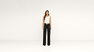 A woman stands against a plain white background, wearing a white sleeveless top tucked into high-waisted, wide-leg black pants with a gold belt, creating an effortlessly chic look.