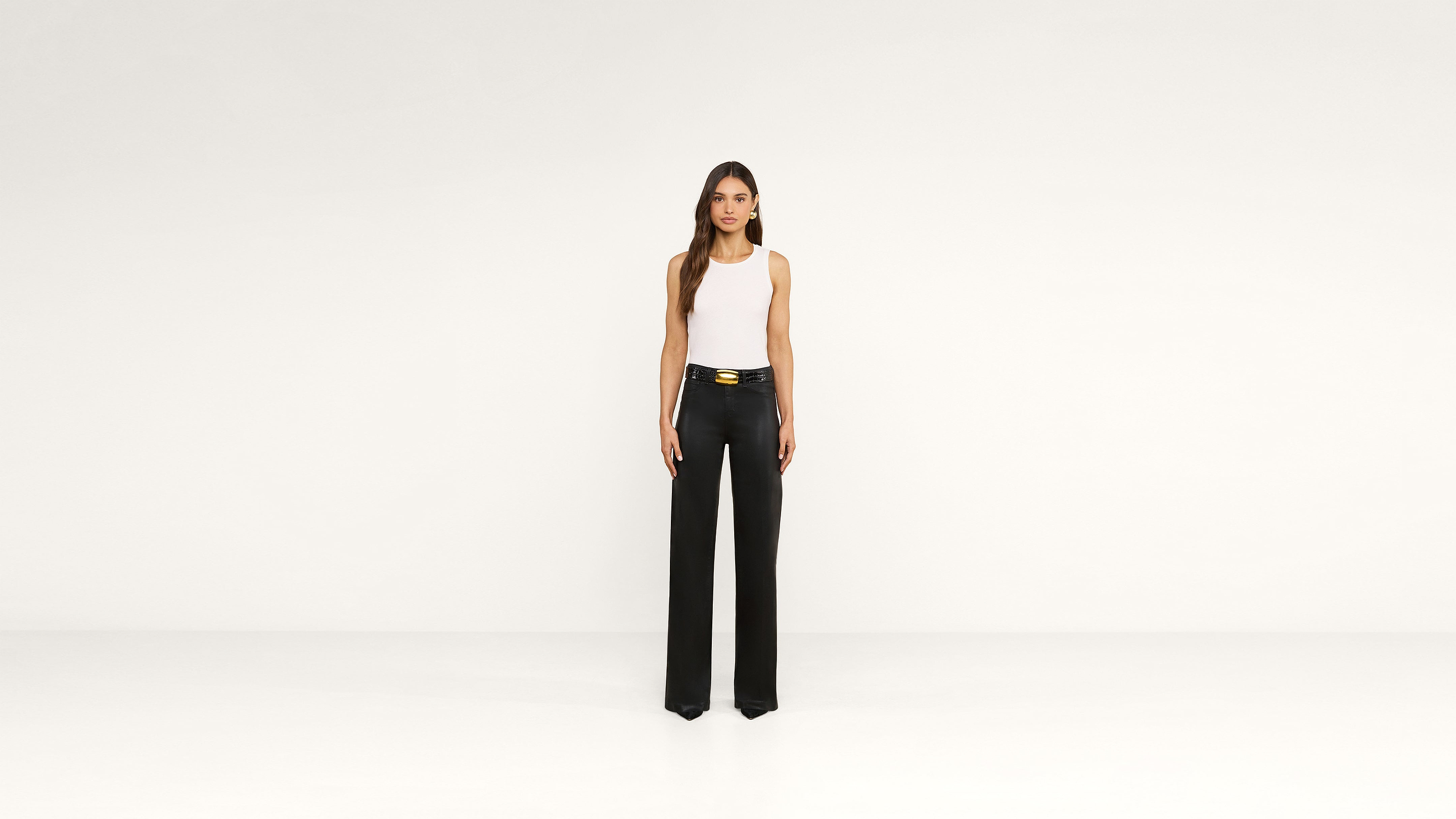 A woman stands against a plain white background, wearing a white sleeveless top tucked into high-waisted, wide-leg black pants with a gold belt, creating an effortlessly chic look.