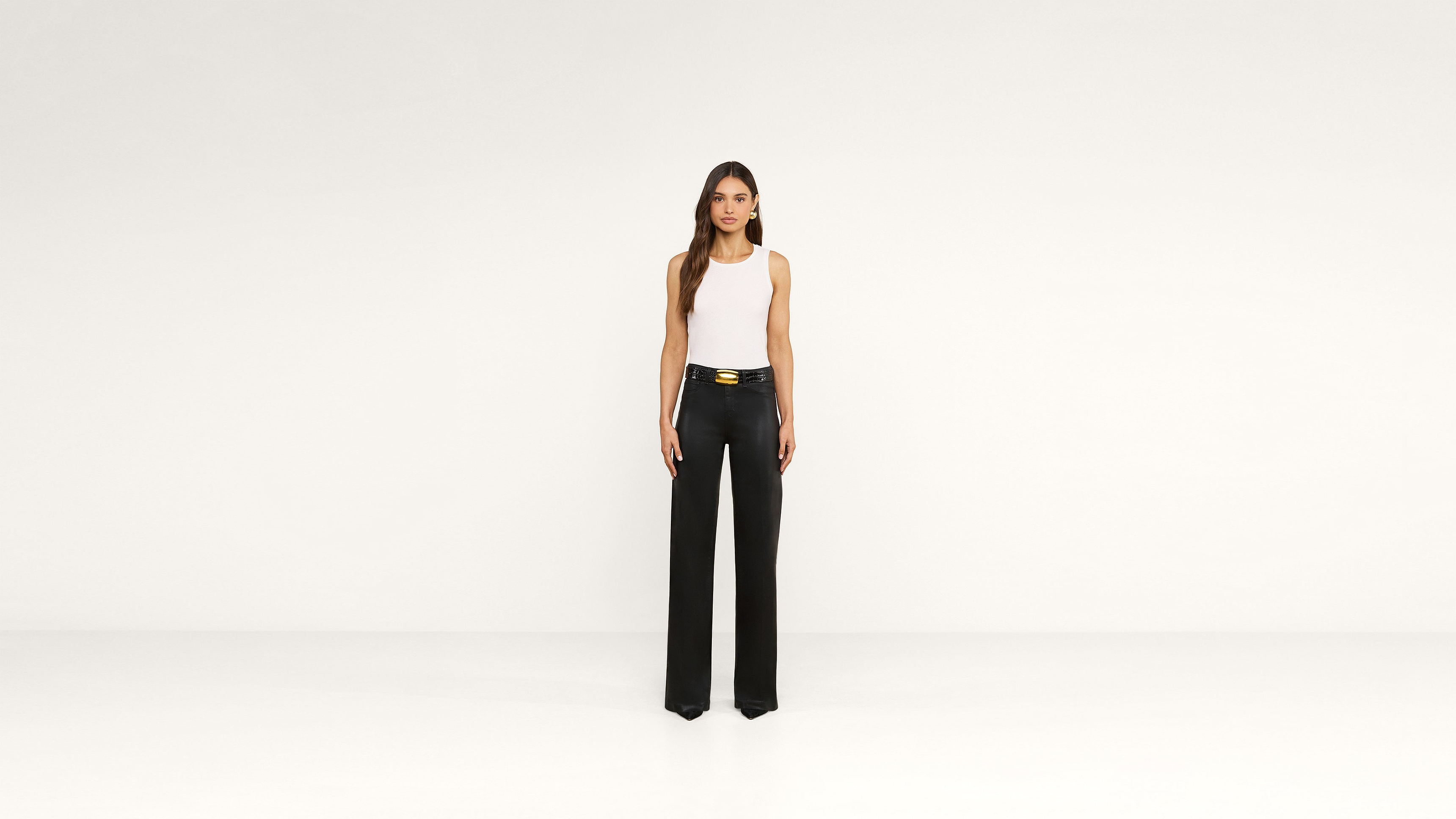 A woman stands against a plain white background, wearing a white sleeveless top tucked into high-waisted, wide-leg black pants with a gold belt, creating an effortlessly chic look.