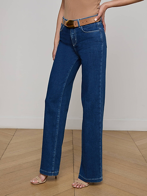 Someone in Clayton Wide-Leg Jeans, indigo-washed and high-rise with a wide-leg fit, paired with a brown belt and tan heeled sandals, stands on a wooden floor against a plain light-colored wall.