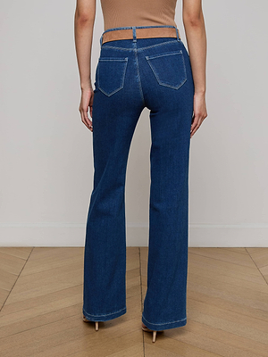 A person stands facing away, wearing the Clayton Wide-Leg Jean in an indigo wash with exposed waistband cutouts, back pockets, and flared legs, paired with a tan top on a wooden floor.