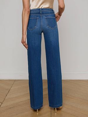 A person stands on a wooden floor wearing the L’AGENCE Clayton Wide-Leg Jean in stretch denim with back pockets and contrast stitching, paired with beige heels. The high-rise, wide-leg jeans are seen from the back.
