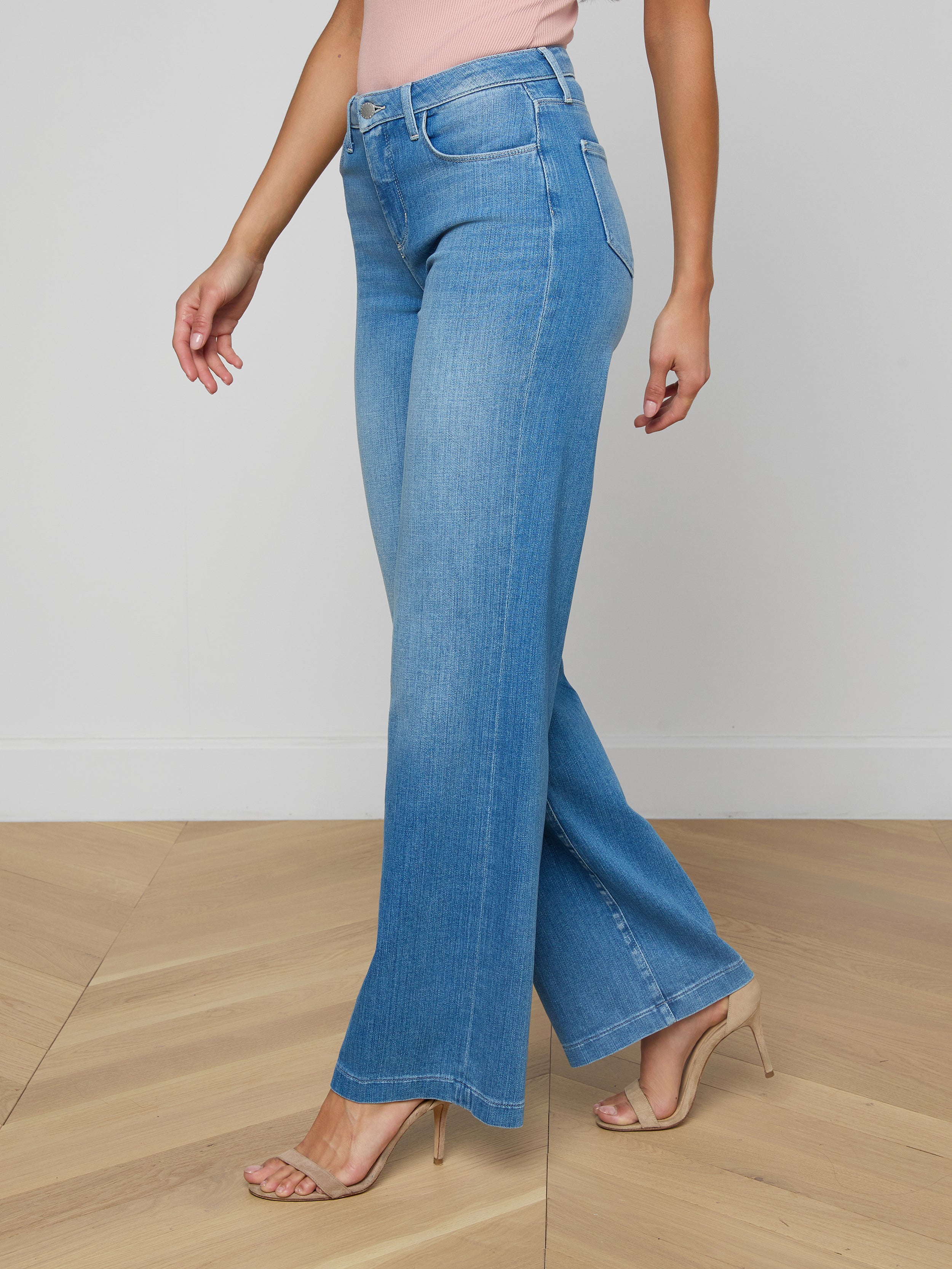 A woman is wearing the Clayton Wide-Leg Jean in light blue, featuring a high-rise and wide-leg silhouette crafted from stretch denim, along with a light pink top and nude high-heeled sandals, as she walks against a plain white background.