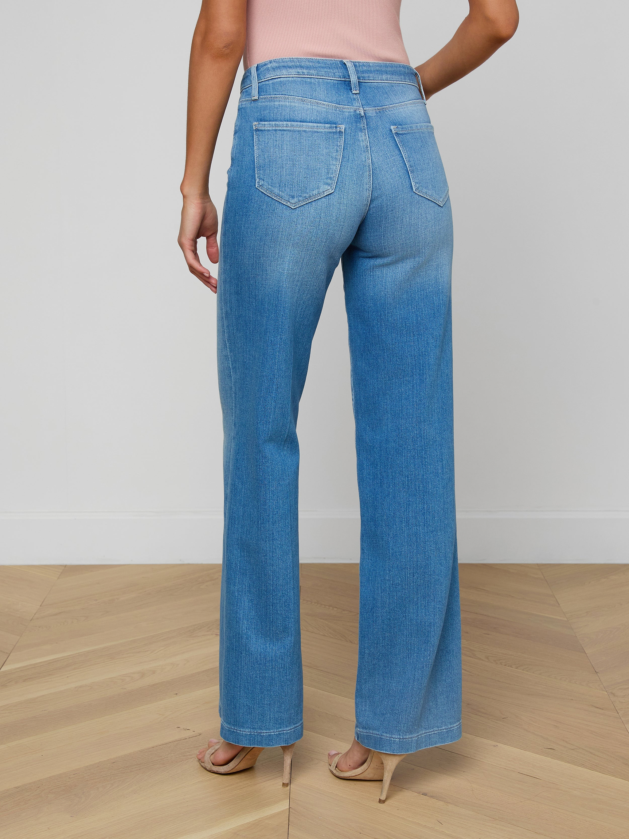 A woman is shown from the back wearing the Clayton Wide-Leg Jean in a light blue high rise style, paired with beige heels and a pink sleeveless top, her left arm bent with her hand on her hip.
