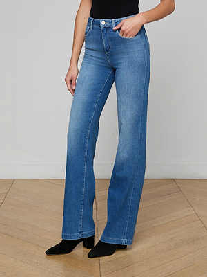 Only the lower half of a person is visible as they stand against a light wall on a wooden floor, wearing the Clayton Wide-Leg Jean in high-rise blue stretch denim, paired with black heeled boots.