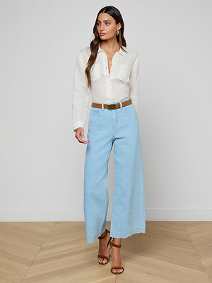 Woman wearing a white blouse and light blue pants with a belt, standing on a wooden floor.