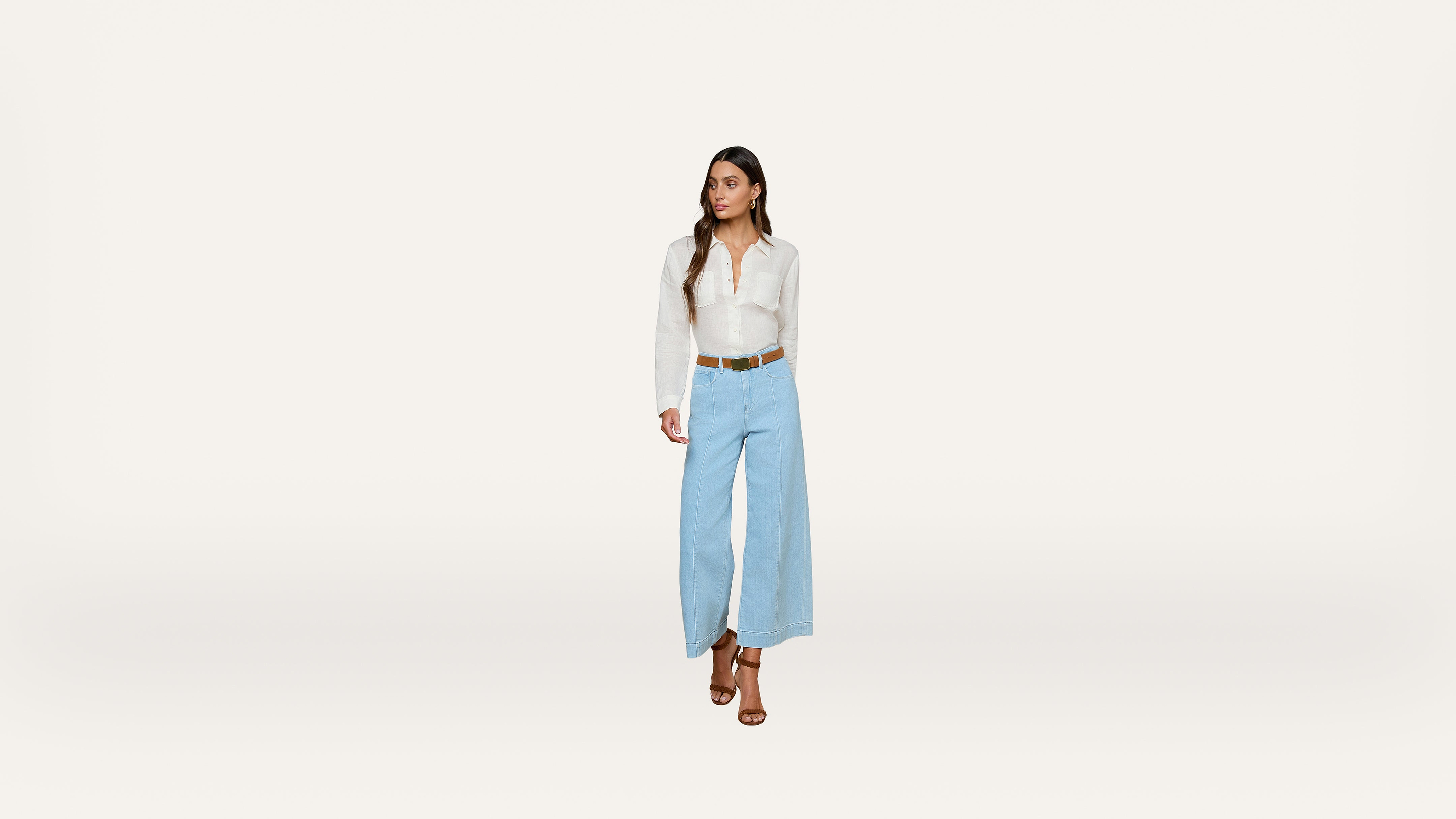 A woman wearing a white long-sleeve shirt, light blue wide-leg pants, a brown belt, and brown heeled sandals stands against a plain white background, looking to her left.