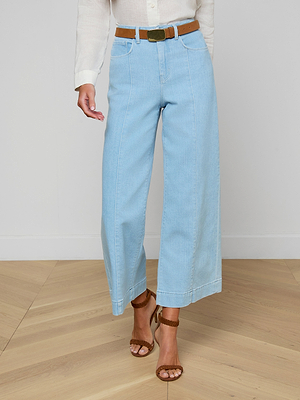 Person wearing light blue jeans with a brown belt and sandals on a wooden floor.